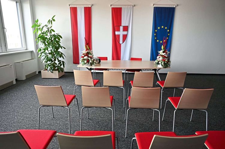 Sessel vor einem Tisch in einem leeren Raum, an dessen Wand die Fahnen von Österreich, Wien und der EU hängen.