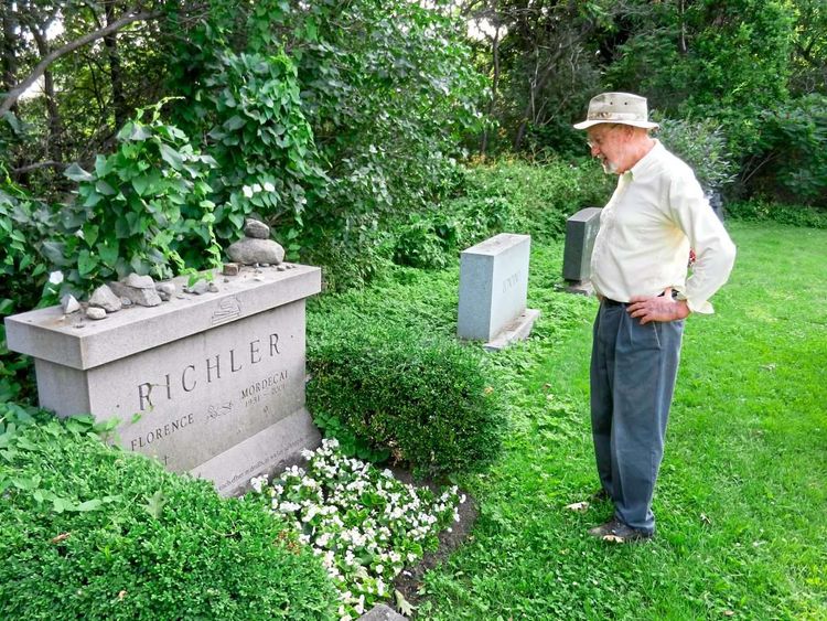 Ein alter Mann mit weißem Hemd und Hut vor einem Grabstein in einer Gartenanlage.