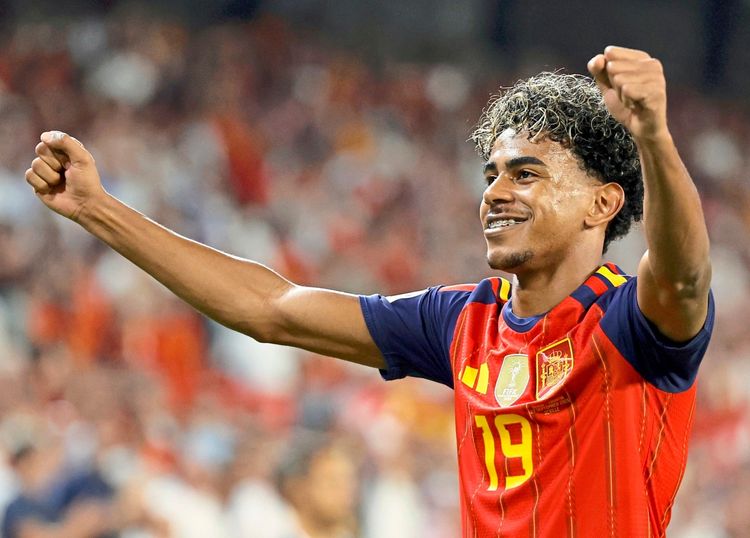 Ein Fußballspieler im roten Trikot der spanischen Nationalmannschaft jubelt mit erhobenen Armen. Im Hintergrund ist ein unscharfes Stadion mit Fans zu sehen.