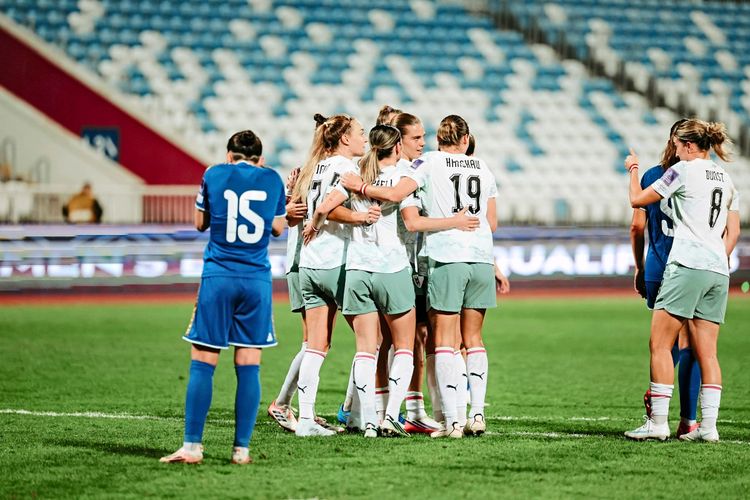 Auf einem Fußballfeld umarmen sich Spielerinnen eines Teams in weißen Trikots mit kurzen Hosen. Eine Spielerin des gegnerischen Teams in einem blauen Trikot steht links im Vordergrund. Im Hintergrund sind leere Ränge eines Stadions zu sehen.