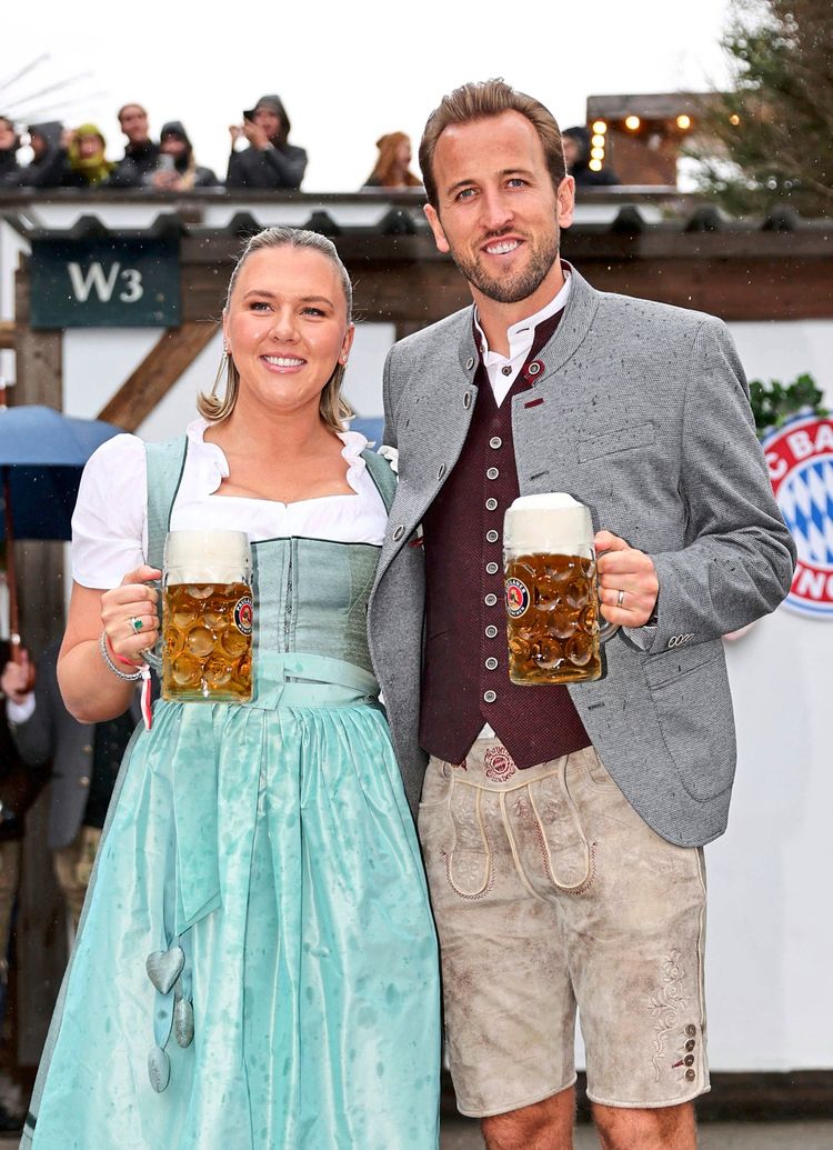 Ein Mann und eine Frau in traditioneller bayerischer Tracht stehen lächelnd nebeneinander. Der Mann trägt eine graue Joppe, ein weißes Hemd und eine beige Lederhose. Die Frau trägt ein mintgrünes Dirndl. Beide halten Krüge mit Bier in den Händen. Im Hintergrund sind weitere Personen und Oktoberfest-Dekorationen sichtbar.