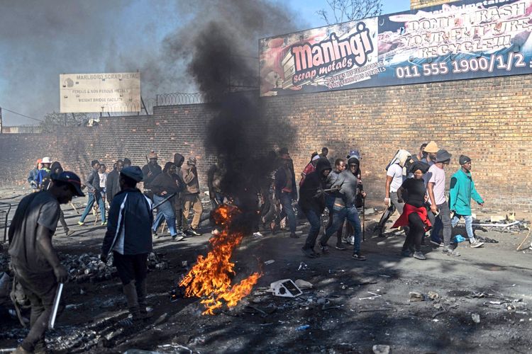 Demonstranten marschieren während eines Anti-Migranten-Protests in Alexandra am 16. Juli 2026. Im Vordergrund brennt ein Feuer, während eine Gruppe von Menschen, teils mit Gegenständen bewaffnet, an einer Straße entlangläuft. Im Hintergrund ist eine Mauer mit Werbeschildern für Altmetall-Recycling zu sehen.