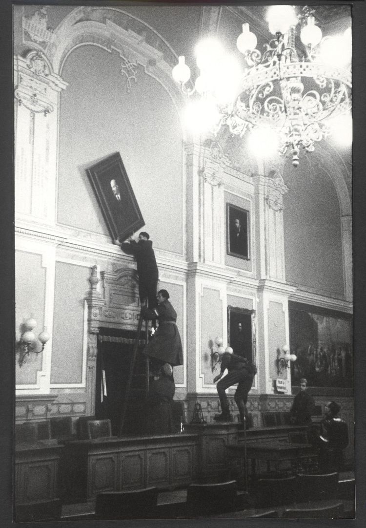Schwarz-weißes Foto von Personen in edlem Gebäude, die ein großes Porträtfoto von der Wand abhängen.
