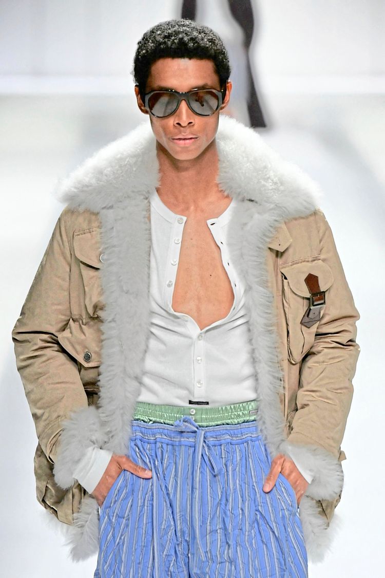 Ein Model trägt eine Modekreation von Dolce & Gabbana aus der Herren-Frühjahr-Sommer-Kollektion 2026 während der Mailänder Fashion Week. Das Outfit besteht aus einer beige Jacke mit weißem Fellkragen, einem weißen Shirt und blauen, gestreiften Hosen. Der Look wird mit einer großen Sonnenbrille abgerundet.