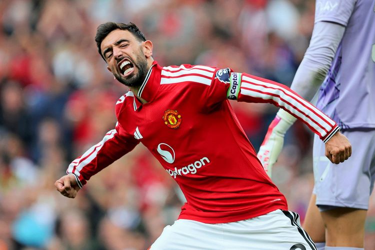 Ein Fußballspieler im roten Trikot von Manchester United jubelt mit ausgebreiteten Armen und einem ausdrucksvollen Gesichtsausdruck während eines Spiels im Stadion Old Trafford. Im Hintergrund sind unscharf andere Spieler und Zuschauer zu erkennen.