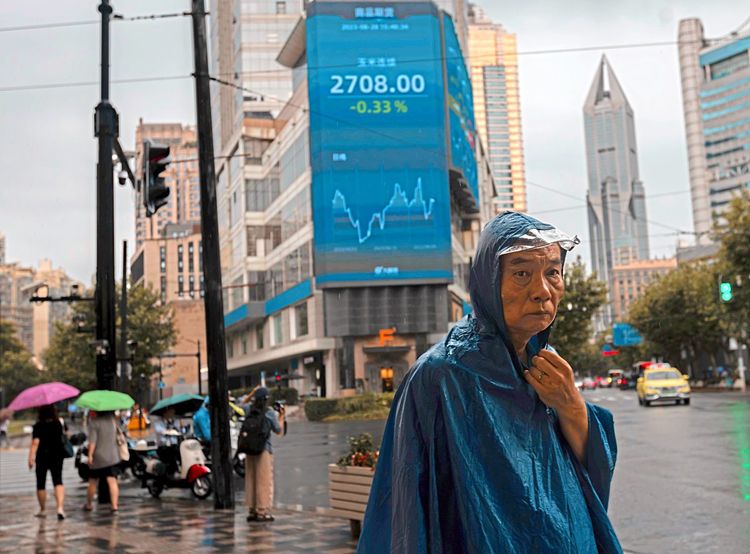 Ein Mann in blauer Regenjacke steht in Schanghai vor einem großen Bildschirm, auf dem die neuesten Börsendaten angezeigt werden.