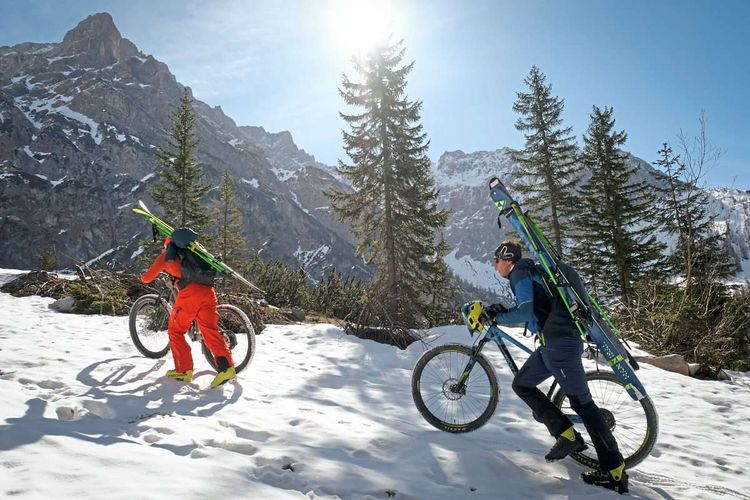 Zwei Menschen die ihre Mountainbikes im Schnee bergauf schieben, am Rücken Tourenski.