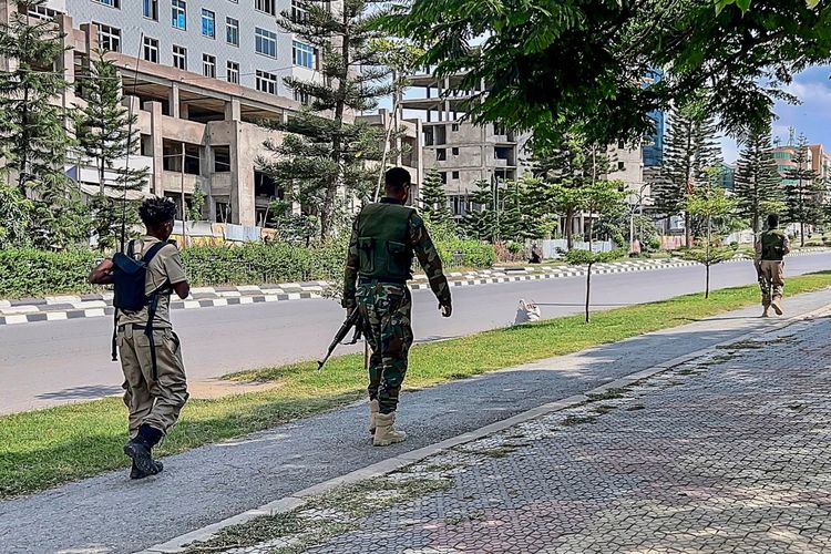 Drei bewaffnete Männer in militärischer Kleidung gehen auf einem Gehweg entlang einer Straße in einer städtischen Umgebung in Mekelle, Äthiopien. Im Hintergrund sind Gebäude, darunter Baustellen, sowie Bäume und eine mehrspurige Straße zu sehen.