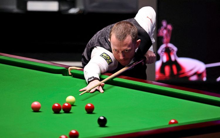 Ein Snooker-Spieler in einem weißen Hemd und einer schwarzen Weste zielt mit einem Queue auf eine Kugel auf einem grünen Snookertisch. Im Hintergrund unscharf beleuchtete Details.