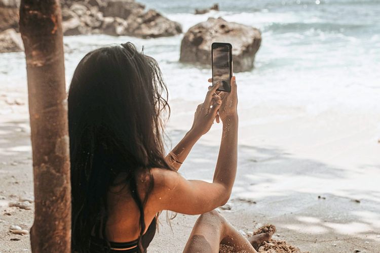Eine junge Frau sitzt am Strand von Bali und fotografiert das Meer mit ihrem Smartphone.