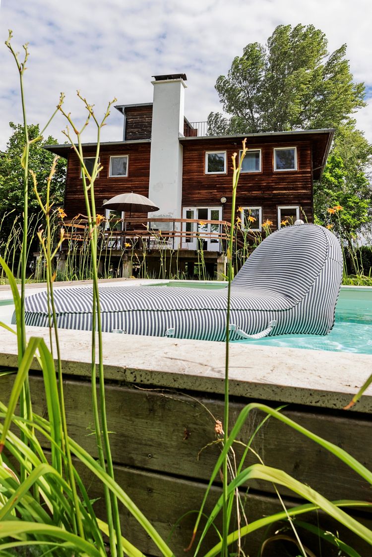 Ein modernes, mehrstöckiges Haus mit Holzfassade, umgeben von Bäumen, im Hintergrund. Im Vordergrund befindet sich ein Pool mit einer gestreiften, schwimmenden Liege. Gräser und Blumen rahmen den unteren Bildrand. Der Himmel ist teilweise bewölkt.