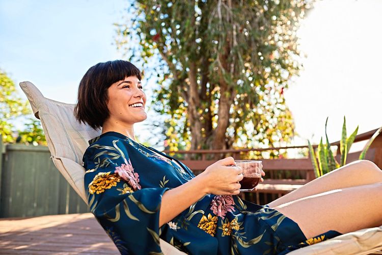 Eine Frau in einem Kimono liegt lächelnd in einem Liegestuhl, eine Tasse Tee in der Hand