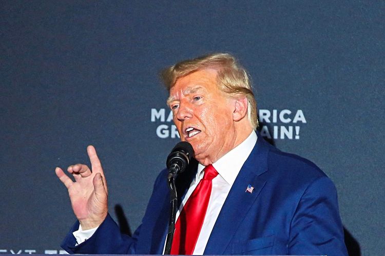 Donald Trump hebt die Hand und redet in ein Mikrofon. Im Hintergrund ist der Schriftzug 