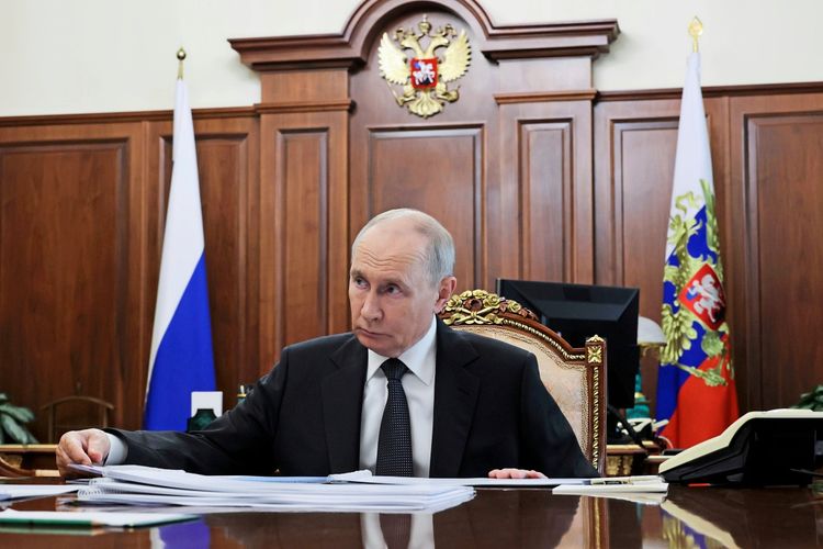 Ein Mann sitzt an einem Schreibtisch in einem prunkvoll eingerichteten Raum mit Holzvertäfelung. Hinter ihm sind die russische Flagge und das russische Staatswappen zu sehen. Vor ihm liegen Dokumente.