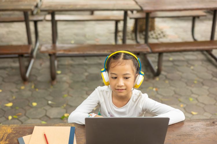 Focused Asian child engages in online learning outdoors, using a laptop and headphones. It shows the role of technology and digital devices in remote education in modern learning environments.
