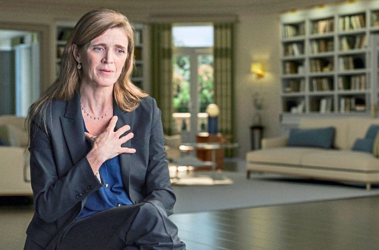 Eine Frau mit langen, bräunlichen Haaren sitzt in einem modernen, hell eingerichteten Wohnzimmer. Sie trägt einen dunkelblauen Blazer und ein blaues Oberteil und hat eine Hand auf ihrer Brust. Im Hintergrund sind ein Bücherregal, ein Sofa und große Fenster mit Blick ins Grüne zu sehen.