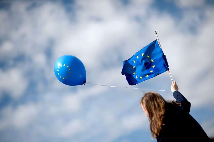 Eine junge Frau hält eine EU-Flagge und einen EU-Luftballon gen Himmel, Rückansicht von unten.