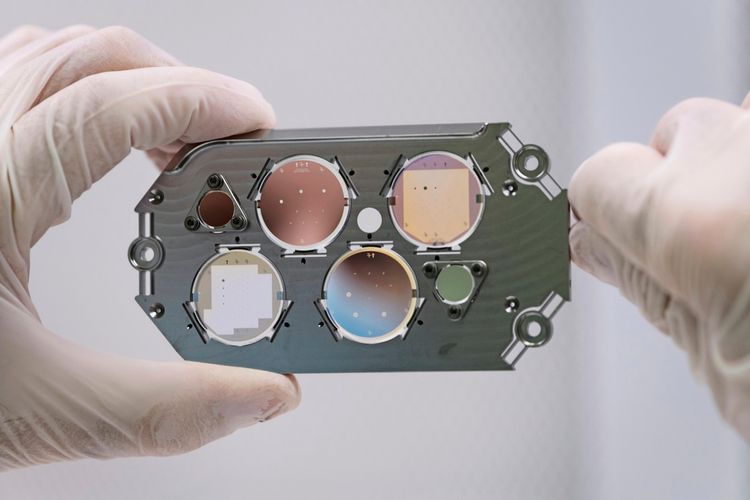 Eine Nahaufnahme einer in Handschuhen gehaltenen Fokalebenenmaske, die zur Lichtunterdrückung und zum Aufspüren von Planeten verwendet wird. Die Maske besitzt mehrere kreisförmige Abschnitte mit fein abgestimmten, lichtundurchlässigen Elementen, die zum Blockieren von Sternlicht dienen.
