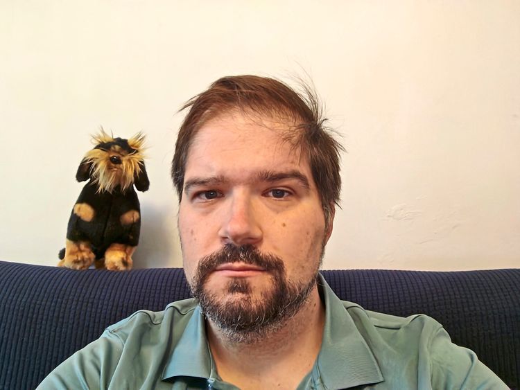 Ein Mann mit Bart und neutralem Gesichtsausdruck sitzt vor einer weißen Wand auf einem blauen Sofa. Hinter ihm steht ein Stofftier in Form eines Hundes mit schwarzem und braunem Fell.