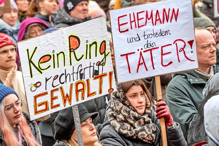 Eine Menschenmenge bei einer Demonstration. Zwei Frauen halten Plakate hoch. Auf dem linken Plakat steht: „Kein Ring rechtfertigt GEWALT!“. Auf dem rechten Plakat steht: „EHEMANN und trotzdem dein TÄTER!“. Die Demonstration findet am Königsplatz in München statt und richtet sich gegen sexualisierte Online-Gewalt.