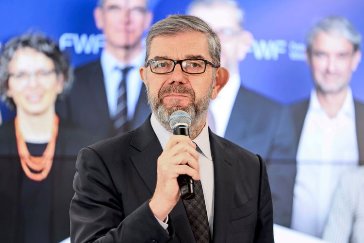 Eine Person in einem schwarzen Anzug und Krawatte hält ein Mikrofon während einer Pressekonferenz. Im Hintergrund befinden sich unscharf abgebildete Personen vor einer blauen Wand mit einem Schriftzug. Die Gesichter sind unkenntlich gemacht.