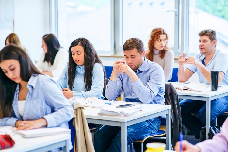 Eine Gruppe von Studierenden sitzt an Tischen in einem Klassenzimmer. Sie arbeiten konzentriert, einige schreiben, lesen oder denken nach. Im Hintergrund sind Fenster und weitere Personen zu sehen.