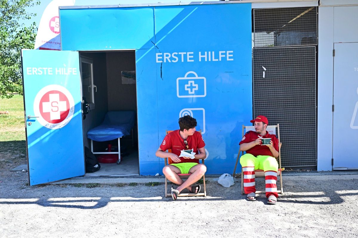 Zwei Rettungssanitäter in roten Shirts und gelb-roten Hosen sitzen vor einer blauen Erste-Hilfe-Station auf Klappstühlen und essen. Hinter ihnen ist die geöffnete Station mit einem Behandlungsbett sichtbar.