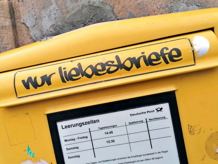 Gelber Briefkasten mit Aufschrift „nur liebesbriefe“ oben. Darunter ein Schild mit Leerungszeiten der Deutschen Post.