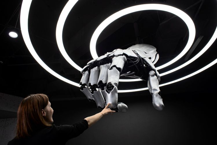 Eine Reporterin berührt eine große, silberne Roboterhand, die von der Decke hängt. Im Hintergrund sind leuchtende Ringe als Dekoration zu sehen. Die Szene befindet sich in der Beijing World of Robots.