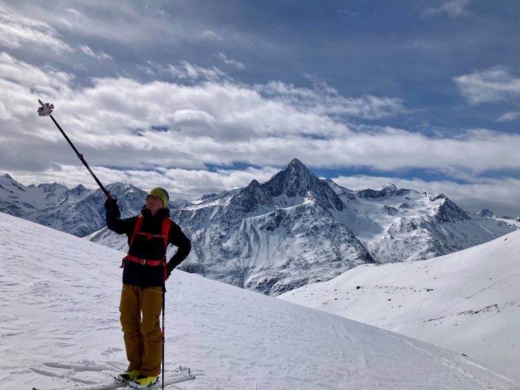 Ein Bergführer steht mit Ski auf der Piste und zeigt mit dem Stock zu einem Berg.