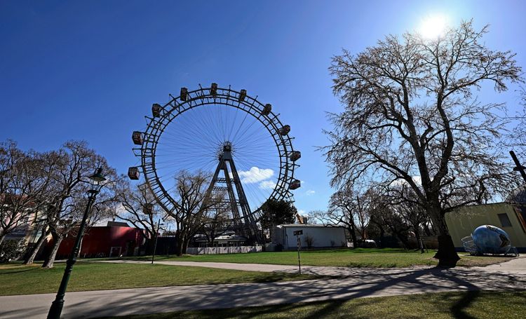Das Bild zeigt das Wiener Riesenrad im Prater an einem sonnigen Tag. Im Vordergrund sind Wiesen, Wege und Bäume, teilweise noch ohne Blätter, zu sehen. Der Himmel ist klar und blau, die Sonne scheint.