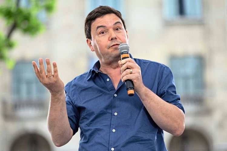 Ein Mann in einem blauen Hemd spricht mit einem Mikrofon in der Hand bei einer Veranstaltung im Freien. Der Hintergrund zeigt ein verschwommenes Gebäude.