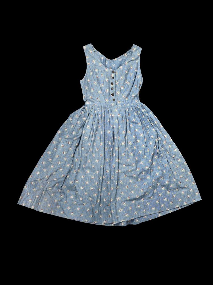 Ein ärmelloses, blaues Kleid mit einem ausgestellten Rock und einem Blumenmuster. Es hat Knöpfe auf der Vorderseite und einen Rundhalsausschnitt. Der Hintergrund ist schwarz.