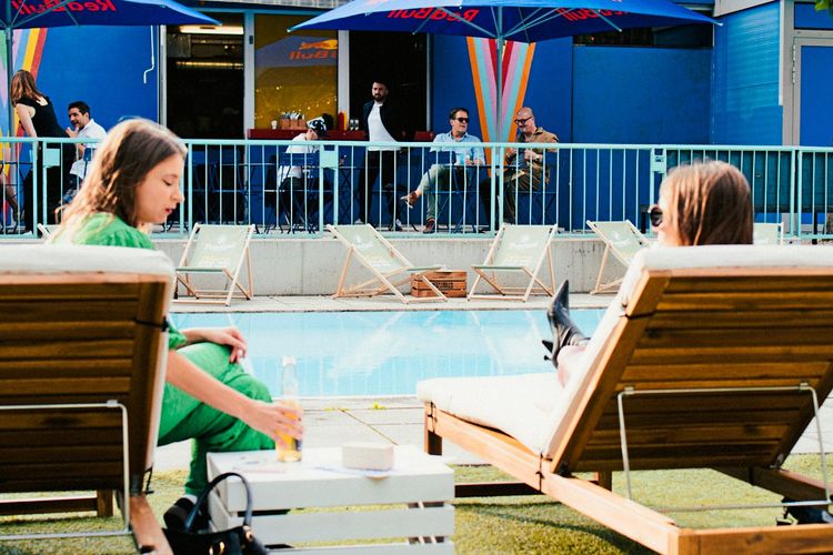 Zwei Frauen entspannen auf Holzliegen am Pool. Eine hält ein Getränk in der Hand. Im Hintergrund sitzen einige Personen auf Stühlen unter Sonnenschirmen, dahinter eine bunte Wand.