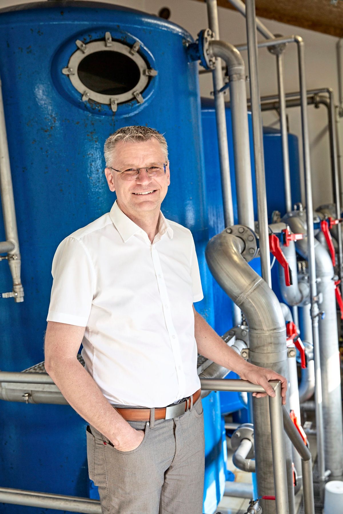 Ein lächelnder Mann in weißem Hemd und grauer Hose steht vor einer großen blauen Industriemaschine mit Metallrohren und Armaturen.