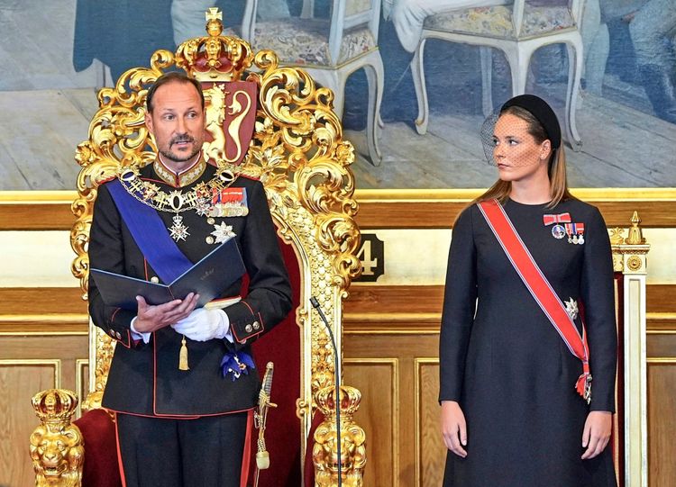 Ein Mann in einer zeremoniellen Uniform sitzt auf einem goldenen Thron mit roten Polstern und liest aus einem Dokument vor, während eine Frau neben ihm steht. Sie trägt ein schlichtes, schwarzes Kleid, einen roten Schärpengürtel mit norwegischer Flagge und Auszeichnungen. Der Hintergrund zeigt eine Wand mit dekorativen Elementen.
