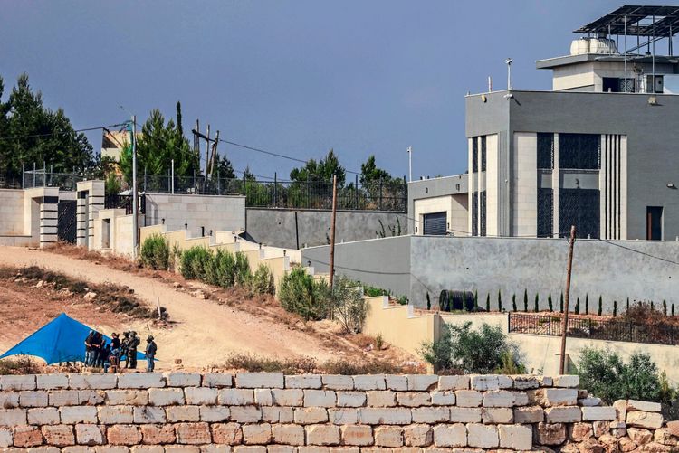 Israelische Soldaten stehen neben einer Gruppe von Personen unter einem blauen Zelt, das außerhalb des Eingangs eines palästinensischen Wohnhauses im Dorf Qusra, südlich von Nablus im israelisch besetzten Westjordanland, aufgestellt wurde. Im Hintergrund ist ein modernes Gebäude mit grauer Fassade und umgebenden Mauern zu sehen. Die Szene findet in einer hügeligen, kargen Landschaft statt.