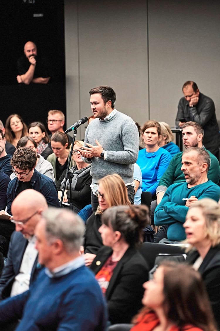 Ein Mann spricht in ein Mikrofon, umgeben von sitzendem Publikum in einem Konferenzraum. Die Anwesenden hören aufmerksam zu oder machen Notizen. Im Hintergrund sitzt ein Mann gedankenverloren mit seiner Hand am Kinn.
