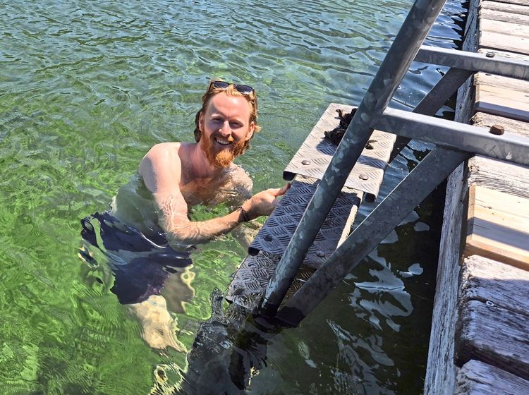 Ein Mann steht bis zur Hüfte im klaren, grünlichen Wasser neben einem hölzernen Steg mit Metallleiter. Er hält eine der Stufen fest und lächelt in die Kamera.