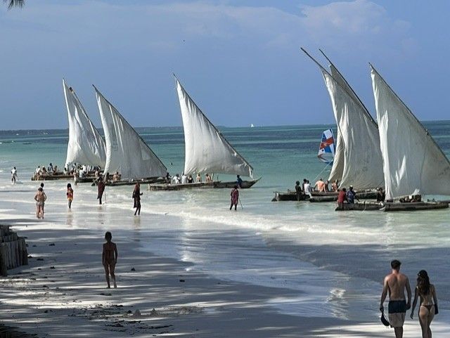 Menschen spazieren am Strand, im Wasser sind Boote mit Segeln 