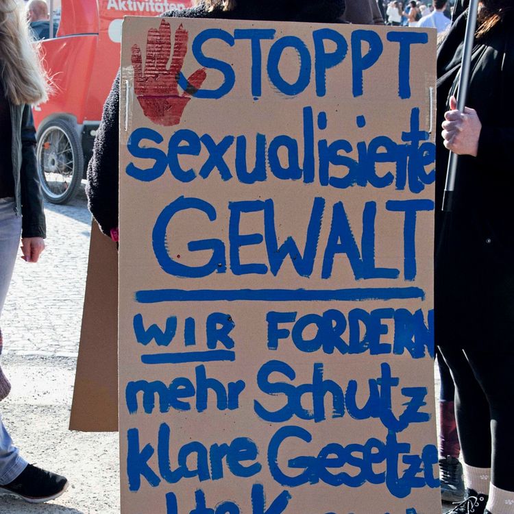 Ein Pappschild bei einer Demonstration zeigt den handschriftlich geschriebenen Text: „Stoppt sexualisierte Gewalt. Wir fordern mehr Schutz, klare Gesetze, echte Konsequenz - online & offline.“ Oben links ist ein roter Handabdruck zu sehen. Im Hintergrund stehen Personen, und es ist ein Teil eines Platzes sichtbar.