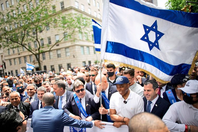 Bei einer Parade in New York City sind viele Menschen zu sehen, darunter einige mit israelischen Flaggen und sichtlich formelle Kleidung tragend. Im Hintergrund befinden sich Gebäude. Die Szene zeigt eine Feierlichkeit oder Demonstration.
