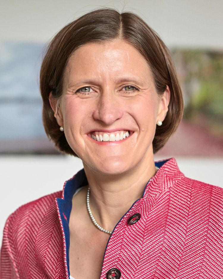 Eine Person trägt eine rosa Jacke mit blauem Kragen und Knöpfen sowie eine Perlenkette. Der Hintergrund zeigt unscharfe Farben und Formen.