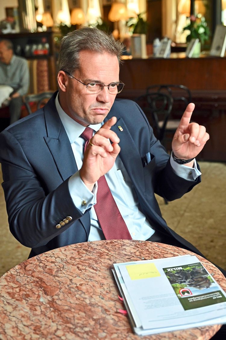 Ein Mann in Anzug und Krawatte sitzt an einem Marmortisch in einem Café. Auf dem Tisch liegen Dokumente und ein Magazin. Er gestikuliert mit erhobenen Händen und schaut ernst. Im Hintergrund ist eine unscharfe Innenraumszene mit weiteren Personen und Dekorationen zu sehen.