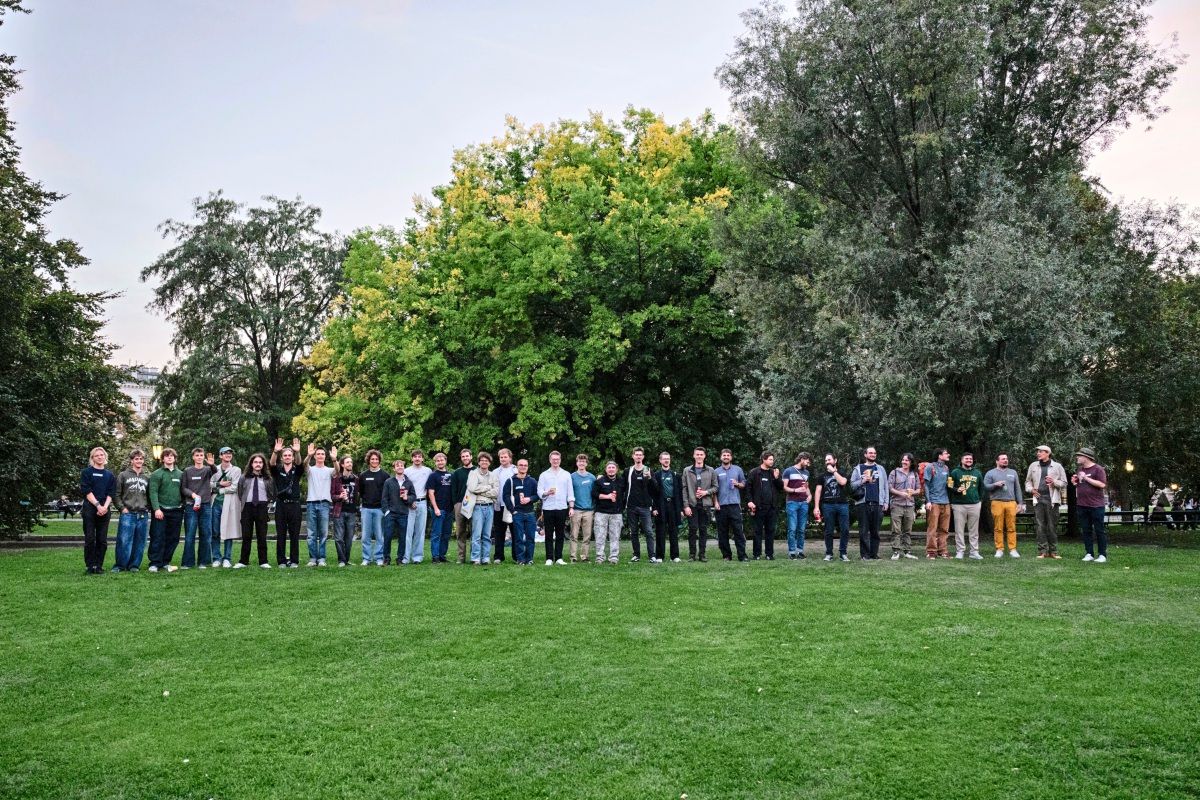 Eine große Gruppe von Menschen posiert auf einer Wiese in einem Park. Im Hintergrund sind Bäume und ein Abendhimmel zu sehen. Die Personen sind von links nach rechts nach Alter aufgestellt.