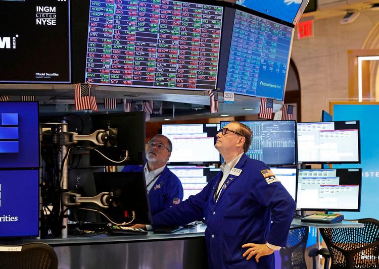 Zwei Händler in blauen Kitteln arbeiten auf dem Parkett der New Yorker Börse. Sie betrachten mehrere Monitore mit Finanzdaten und Börsentickern. Kleine US-Flaggen schmücken die Handelsstation.