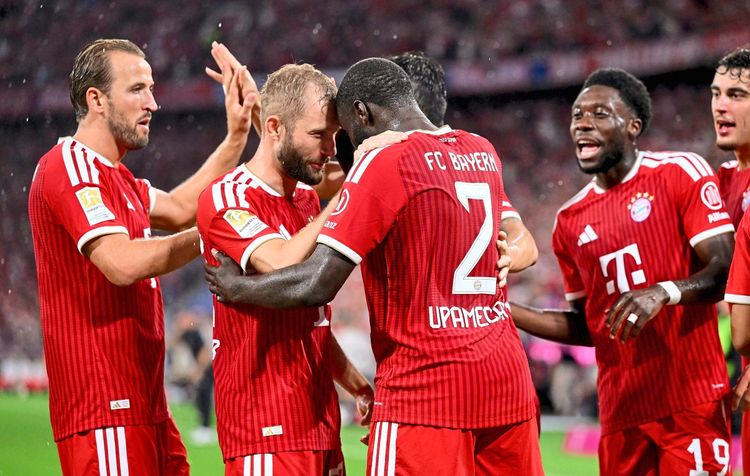 Mehrere Spieler des FC Bayern München in roten Trikots jubeln gemeinsam nach einem Tor. Dayot Upamecano steht im Mittelpunkt und wird von seinen Mitspielern, darunter Konrad Laimer, Harry Kane und Alphonso Davies, umarmt und gefeiert. Im Hintergrund sind weitere Spieler und das Stadionpublikum zu sehen.