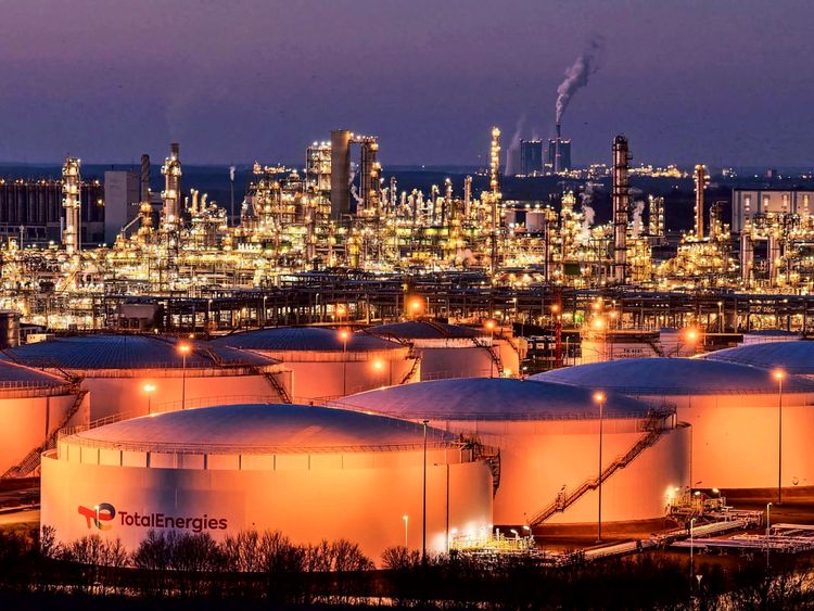 Blick auf die beleuchteten Anlagen der Total Energies Raffinerie im Chemiepark Leuna bei Nacht. Im Vordergrund befinden sich mehrere große, runde Tanks, während im Hintergrund zahlreiche industrielle Strukturen und das Kraftwerk Schkopau zu sehen sind.