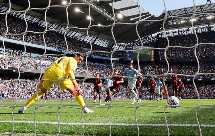 Ein Fußballspiel im Etihad Stadium, Manchester. Ein Tor wird aus der Perspektive hinter dem Netz festgehalten. Ein Spieler im gelben Torwarttrikot schaut auf den Ball, der gerade die Torlinie überquert. Im Hintergrund jubeln Spieler und Zuschauer.