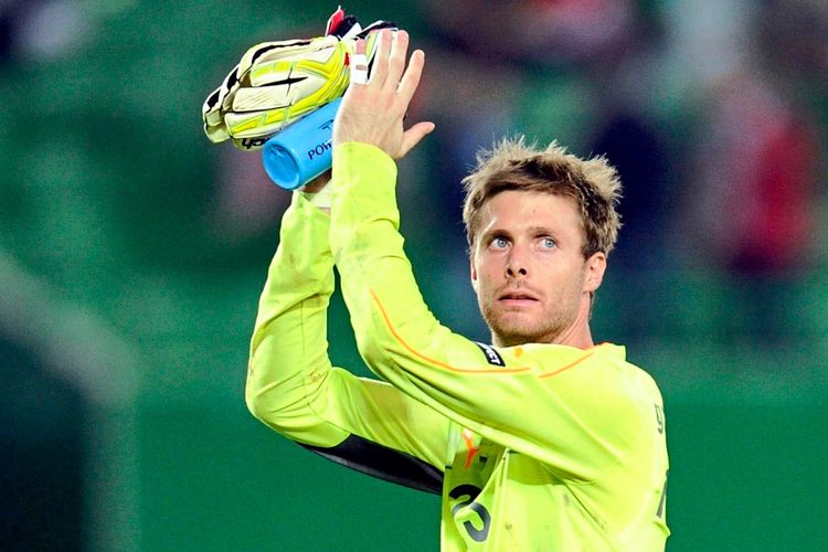 Ein Fußballtorwart in gelber Spielkleidung klatscht mit den Handschuhen über dem Kopf. Der Hintergrund zeigt ein unscharfes Stadion.